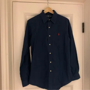 Ralph lauren skjorta - Säljer en klassisk och stilren mörkblå skjorta från Ralph Lauren i storlek S. Skjortan är i mycket gott skick och har endast använts ett fåtal gånger. Den är tillverkad i 100 % bomull med en bekväm passform som passar både till vardag och mer uppklädda tillfällen.  Den ikoniska röda Polo-loggan är broderad på bröstet och knapparna är i vitt för en snygg kontrast mot det mörkblå tyget.