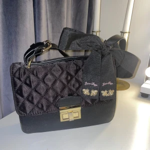 Svart axelremsväska med rosett - Svart axelväska från Call It Spring med quiltat mönster och stor dekorativ rosett. Väskan har guldfärgat spänne och avtagbar axelrem. Detaljer med broderade bin och texten 'Love Bee' framtill. Perfekt för att lyfta din outfit. Har en innerficka och man får med ett extra band. Rosetten är avtagbar.