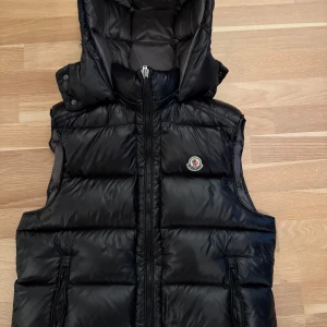 Svart dunväst från Moncler - Säljer en svart dunväst från Moncler med huva och dragkedja framtill. Västen har Moncler-logga på bröstet och två fickor med dragkedja. Levereras med originalpåse och etiketter.