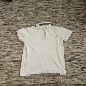 Vit piké från Tommy Hilfiger - Säljer en klassisk vit pikétröja från Tommy Hilfiger med liten broderad logga på bröstet. Tröjan har korta ärmar, krage och knappar vid halsen. Perfekt för en stilren och avslappnad look.