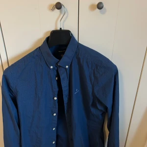 Gant skjorta - Säljer en blå skjorta från GANT med små vita prickar och klassisk button-down krage. Skjortan har långa ärmar och knäppning framtill. Perfekt för dig som gillar stilrena detaljer och snygg passform.