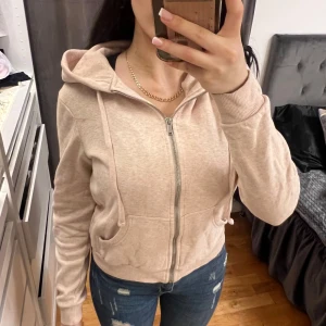 Beige hoodie med dragkedja från H&M - Beige kofta från H&M med dragkedja framtill och huva med dragsko. Tröjan har långa ärmar, ribbade muddar och fickor på magen. Perfekt för en avslappnad och bekväm stil. Storlek Xs från H&M.