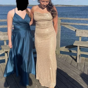 Glittrig guldig långklänning från Stylecheat - Säljer en glittrig guldig långklänning från Stylecheat med smala axelband och figurnära passform. Klänningen har en elegant, skimrande yta och är perfekt för festliga tillfällen. Dragkedja i ryggen för enkel påklädning.