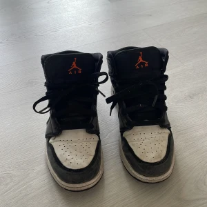Nike Air Jordan 1 Mid svart/vit/blå - Säljer ett par Nike Air Jordan 1 Mid i svart, vit och blå med orange Jumpman-logga på plösen. Skorna har klassisk snörning, rund tå och platt sula. Perfekta för dig som gillar streetwear och vill sticka ut med en ikonisk sneaker.
