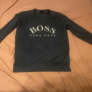 Mörkblå sweatshirt från Hugo Boss - Säljer en mörkblå sweatshirt från Hugo Boss med stor vit logga på bröstet. Tröjan har rund halsringning och långa ärmar. Klassisk och stilren design med ribbade muddar vid ärmslut och nederkant. Priset kan diskuteras! Va inte rädd med att ställa frågor.