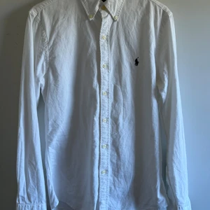 Vit skjorta från Ralph Lauren - Klassisk vit skjorta från Ralph Lauren med broderad logga på bröstet. Slim fit-modell med långa ärmar och knappar framtill. Perfekt för en stilren och clean look. Mycket bra skick. Storlek s