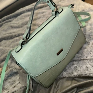 Mintgrön axelväska med strukturdetaljer - Säljer en stilren mintgrön axelväska med både handtag och axelrem. Väskan har en snygg tvåfärgad design med ljusgrön nederdel i strukturmönster och flera fack med dragkedja inuti. Perfekt för att bära dina viktigaste saker.