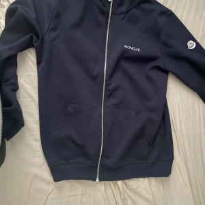 Mörkblå zip-jacka från Moncler - Tja, säljer en stilren mörkblå zip up från Moncler med dragkedja framtill och två fickor. Jackan har lång ärm, Moncler-logga på bröstet och ärmen samt ribbade muddar. Perfekt för dig som gillar klassisk och sportig stil. Den är använd endast en gång och varit i garderoben resten av tiden. Pris kan diskuteras!