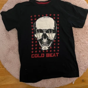 Svart t-shirt med dödskalletryck och röda stjärnor - Cool svart t-shirt med stort dödskalletryck och röda stjärnor på framsidan. Texten 'COLD BEAT' i rött under motivet. Rund halsringning med röd insida. Perfekt för dig som gillar en edgy stil.
