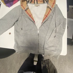 Grå hoodie med dragkedja från Burberry - Säljer en grå hoodie från Burberry med klassiskt rutigt foder i huvan och broderad logga på bröstet. Tröjan har dragkedja, fickor och justerbar huva med snören. Perfekt för en stilren och bekväm look. Pris diskuteras