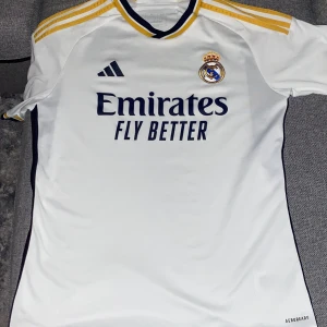 Real Madrid tröja 23/24 - Säljer en vit Real Madrid fotbollströja från Adidas med gula och svarta detaljer. Tröjan har korta ärmar, klubbmärke och Adidas-logga på bröstet samt 'Emirates Fly Better' tryckt på framsidan. Klassisk v-ringad krage med gula och svarta ränder.