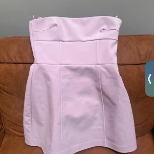 Ljusrosa axelbandslös klänning med rosett - Supersöt ljusrosa axelbandslös klänning med stor rosett bak. Klänningen har en figurnära överdel och utsvängd kjol, perfekt för speciella tillfällen. Dragkedja i sidan för enkel påklädning.