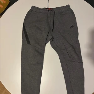 Grå mjukisbyxor från Nike - Säljer ett par grå mjukisbyxor från Nike med snörning i midjan och diskret logga på benet. Byxorna har en bekväm passform och är perfekta för en avslappnad stil. De har också praktiska fickor med dragkedja.