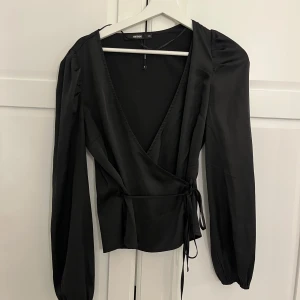 Svart omlottopp från Bik Bok - Originalpris: 500kr . Säljer en svart omlottopp från Bik Bok med långa ärmar och knyt i midjan. Toppen har en djup v-ringning och är tillverkad i ett glansigt, mjukt material som ger en elegant känsla.