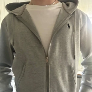 Grå hoodie från Ralph Lauren - Säljer en stilren grå hoodie från Ralph Lauren med dragkedja och huva. Tröjan har fickor framtill och den klassiska loggan broderad på bröstet. Perfekt för en avslappnad look. 