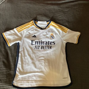 Real Madrid hemmatröja 23/24 från Adidas - Säljer en vit Real Madrid fotbollströja från Adidas med gula detaljer och svarta inslag. Tröjan har klubbmärke och sponsortryck på bröstet samt texten 'Hala Madrid' i nacken. Klassisk passform och korta ärmar. Bara använde några få gånger. Pris kan diskuteras.