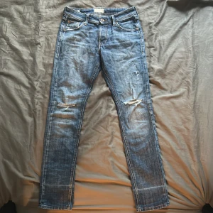 Blå slitna jeans med raka ben - Säljer ett par blå jeans med slitningar på knäna och lätt tvättad look. Jeansen har raka ben, klassiska fem fickor och gylf med dragkedja och knapp. Storlek W29 L32. Perfekta för en avslappnad stil.