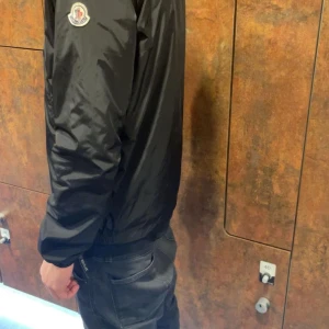 Svart vindjacka från Moncler windbreaker - Säljer en svart vindjacka från Moncler med huva och dragkedja. Jackan har Moncler-logga på ärmen och en snygg randig detalj i rött, vitt och blått på huvan. Perfekt för dig som vill ha en stilren och sportig look. Storlek S/1 i bra skick 9/10. Kan byta mot moncler cardigan eller windbreaker i storlek M/L svart