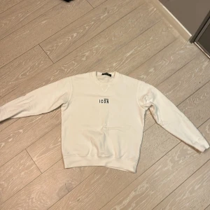 Vit sweatshirt från Dsquared2 med 'ICON'-tryck - Säljer en vit sweatshirt från Dsquared2 med svart 'ICON'-tryck på bröstet. Tröjan har rund halsringning och långa ärmar. Perfekt för en clean och stilren look.