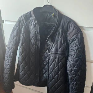 Svart quiltad jacka från Replay - Säljer en svart quiltad jacka från Replay med dragkedja och en bröstficka med zip. Jackan har lång ärm och diskret Replay-logga på ärmen. Snygg, lätt och passar perfekt till kyligare dagar.