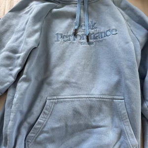 Ljusblå hoodie från Peak Performance - Säljer en ljusblå hoodie från Peak Performance med broderad logga på bröstet. Tröjan har huva med dragsko och en stor magficka framtill. Perfekt för en avslappnad stil.