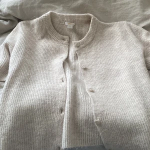 Beige stickad kofta från Gina Tricot - Mysig beige stickad kofta från Gina Tricot med rund halsringning och knappar framtill. Koftan har långa ärmar och ribbad struktur, perfekt för lager-på-lager under kyliga dagar.