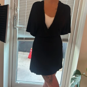 Svart omlottklänning med kort ärm - Säljer en svart omlottklänning med kort ärm och v-ringning. Klänningen har en figurnära passform och är i kort modell, perfekt för dig som gillar en stilren look. Materialet känns mjukt och följsamt mot kroppen. 