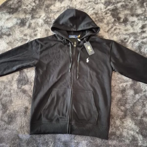 Svart hoodie från Polo Ralph Lauren - Svart hoodie från Polo Ralph Lauren med dragkedja framtill och klassisk logga broderad på bröstet. Tröjan har huva med dragsko och två fickor framtill. Perfekt för en avslappnad och stilren look.