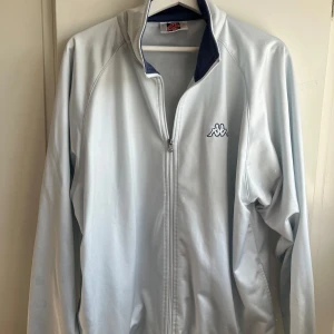 Ljusblå träningsjacka från Kappa - Säljer en ljusblå träningsjacka från Kappa med dragkedja framtill och klassisk logga på bröstet. Jackan har lång ärm och hög krage med mörkblå insida. Perfekt för sportiga outfits eller som ett snyggt lager. - Tröjan har fläckar på ryggen som inte syns jättetydligt men man ser det på bild 3.