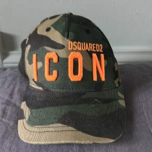 Camo keps från Dsquared2 ICON - Tja! Säljer en camouflage färgad keps från Dsquared2 med den orange texten ICON framtill och 'Dean & Dan Caten' baktill. Kepsen har böjd skärm och justerbar passform. Perfekt för dig som gillar streetwear och vill sticka ut.