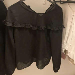 Svart blus med volang från ONLY - Säljer en svart blus från ONLY i storlek 38. Blusen har långa ärmar och en volangdetalj som går runt överdelen. Perfekt för dig som gillar en stilren look med en liten twist.