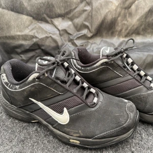 Svarta Nike sneakers med vit swoosh - Svarta sneakers från Nike med vit swoosh på sidan och svarta skosnören. Skorna har meshpaneler för extra andningsförmåga och en platt sula. Perfekta för vardagsbruk och sportiga outfits.