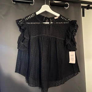 Svart blus med spets och volang från Nelly - Säljer en svart blus från Nelly med transparent spets, volangdetaljer på axlarna och knytning i nacken. Blusen har korta ärmar och ett lätt, luftigt tyg med diskret randigt mönster. Perfekt för dig som gillar romantiska detaljer.