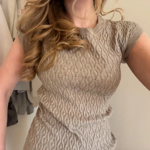 Beige stickad kortärmad topp - Säljer en beige stickad topp med korta ärmar och ett snyggt vågigt mönster. Tröjan har ribbade kanter vid hals, ärmslut och nederkant samt en liten slits i sidan. Perfekt för lager-på-lager eller sommarens outfits.