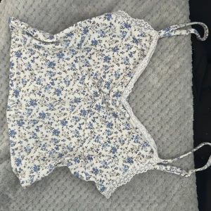 Vit blommig topp med spets - Supersöt vit linnetopp med blått blommönster och tunna axelband. Toppen har spetsdetaljer längs kanterna och en liten rynkning framtill. Perfekt för varma dagar och enkel att matcha med jeans eller kjol. Köpt secondhand så vet ej märke eller storlek, men passar S💞