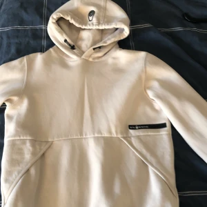 Beige hoodie från Sail Racing - Säljer en stilren beige hoodie från Sail Racing med logga på bröstet och huvan. Tröjan har en klassisk passform, lång ärm och praktisk huva med dragsko. Perfekt för en avslappnad och sportig look.