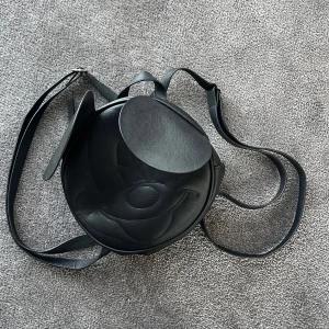 Svart rund ryggsäck med öron - Säljer en svart rund ryggsäck i syntetmaterial med justerbara axelband. Väskan har ett Musse pigg ansikte. Använd ett fåtal gånger.
