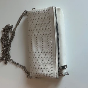 Vit axelväska med nitar och kedja - Säljer en vit axelväska med silverfärgade nitar på framsidan och en grov silverkedja som axelrem. Väskan har dragkedja upptill och är perfekt för att bära det viktigaste. Snygg och trendig accessoar!