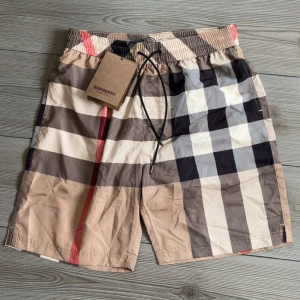 Burberry rutiga badshorts - Snygga badshorts från Burberry med klassiskt rutigt mönster i beige, svart, vitt och rött. Shortsen har elastisk midja med snörning och innerfoder i mesh. Perfekta för sommaren och strandhäng. ‼️Dessa är 100% äkta‼️