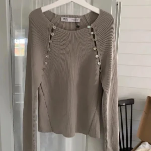 Beige ribbad tröja med knappar från Zara - Säljer en beige ribbad tröja från Zara med långa ärmar och dekorativa knappar längs axlarna. Tröjan har en rund halsringning och är perfekt för lager-på-lager-stil. Snygg och enkel att matcha till olika outfits.