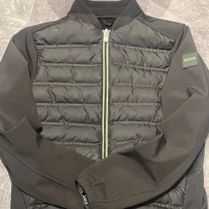 Svart cardigan från HUGO BOSS - Svart cardigan från BOSS med quiltade paneler framtill och släta ärmar. Jackan har dragkedja och en BOSS-logga på ärmen. Perfekt för kyliga sommar dagar och har en modern, sportig look.