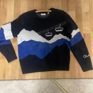 Mörkblå stickad tröja med bergsmotiv från GANT - Säljer en mörkblå stickad tröja från GANT med coolt bergsmotiv i blått, vitt och svart samt två linbanor på framsidan. Tröjan har rund halsringning och långa ärmar, med GANT-logga broderad på ena ärmen.