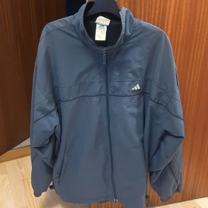 Blå vindjacka från Adidas - Säljer en blå vindjacka från Adidas med dragkedja framtill och två fickor. Jackan har hög krage och Adidas-logga på bröstet. Perfekt för svala dagar och sportiga outfits. Oversize passform