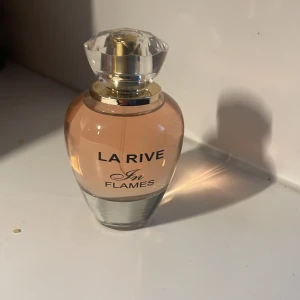 La Rive In Flames parfym - Säljer en elegant parfymflaska från La Rive med doften In Flames. Flaskan är rundad med en transparent glaskropp och ett persikofärgat innehåll. Locket är kristalliknande och silvrigt, vilket ger en lyxig känsla.