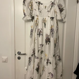 Vit blommig långklänning med puffärmar - Superfin långklänning i vit bomull med blommigt mönster i lila, gult och grönt. Klänningen har puffiga korta ärmar, markerad midja och volang nertill. Perfekt för sommarens alla tillfällen! passar storlek M-L bra