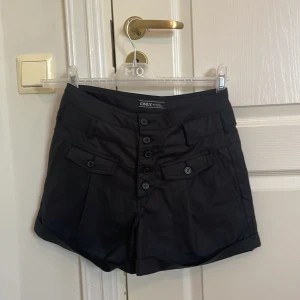 Svarta shorts från ONLY med knappar - Svarta shorts från ONLY med flera dekorativa knappar framtill och fickor både fram och bak. Klassisk design med uppvikta benslut och bälteshällor. Perfekta för en stilren och enkel look.