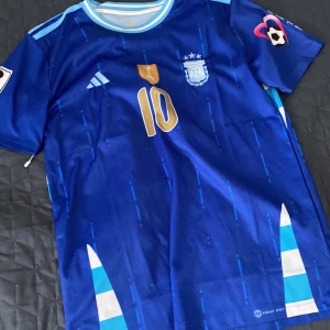 Argentina bortatröja Messi 10 VM 2022 - Säljer en blå Argentina bortatröja från Adidas med Messi 10-tryck i guld. Tröjan har korta ärmar, vita och ljusblå detaljer samt VM-märke på bröstet. Perfekt för fotbollsfans som vill stötta sitt lag.