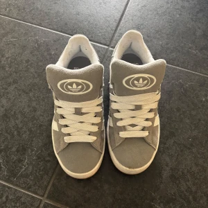Grå Adidas sneakers med vita detaljer - Säljer ett par grå Adidas sneakers i mocka med vita ränder och logga på plösen. Skorna har vita snören och en ljus sula. Perfekta för dig som gillar klassisk streetstil och vill ha bekväma skor till vardags.