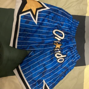 Blå Orlando Magic basketshorts från Just Don - Säljer ett par blå basketshorts från Just Don med Orlando Magic-tryck, vita ränder och stora stjärndetaljer i guld och svart. Shortsen har resår i midjan och snörning. Perfekta för dig som gillar sportig stil.