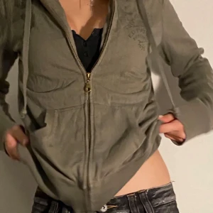 Olivgrön hoodie med dragkedja från Ed Hardy - Säljer en olivgrön hoodie från Ed Hardy med dragkedja och huva. Framsidan har diskret tryck och baksidan har en stor färgglad ros och fjärilar. Mjukt material och ribbade muddar. Perfekt för en avslappnad stil.